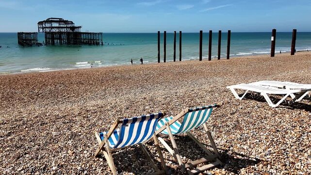 Blue and white striped deck chairs lining Brighton Beach, with the iconic remains of the West Pier standing in the distance beneath a bright coastal sky. Pull back shot.