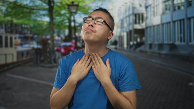 Young chinese man in blue t shirt clutching neck and grimacing on a tree lined city street near canal and parked bicycles; discomfort.