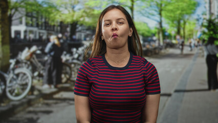 Fototapeta premium Woman makes fish face facing camera on city street with parked bicycles and leafy trees visible; playful mood.