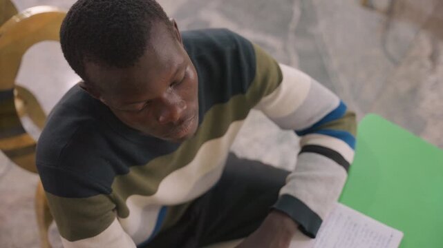 Black man writing love letter at table, young black student scribbling heartfelt note in green notebook, sitting on wooden chair, pen pauses, nervous smile, preparing date surprise, warm soft light,