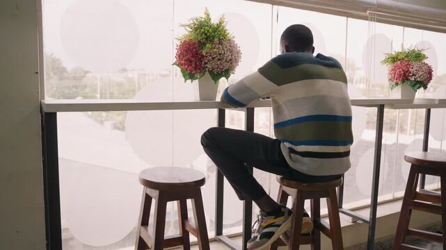 Man at sunlit cafe window. Solitary man in striped sweater sits on wooden stool, gazing through patterned curtain at city street, bouquet on counter beside him, phone untouched, hands folded, quiet