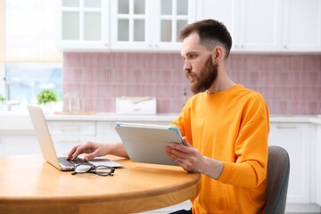 Fototapeta premium Man using tablet while working with laptop at wooden table in kitchen