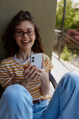 Obraz premium Cheerful woman at home, sitting on a balcony, smiling with phone in hand, striped tee, relaxed posture, sunlit moment, natural colors, contemporary lifestyle