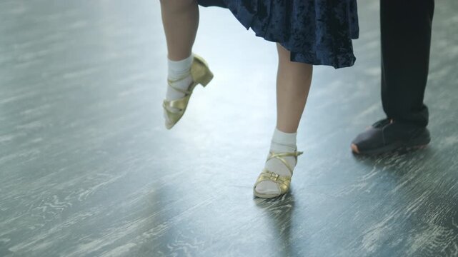 Child dancer's feet in golden high heel shoes training on a parquet floor during choreography lesson.