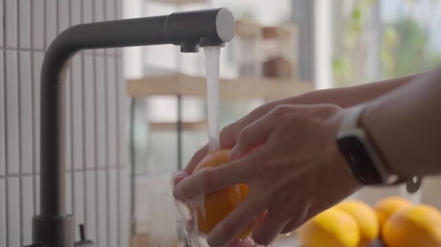 Hands wash orange under running water from kitchen faucet. Concept of fruit cleaning and food hygiene