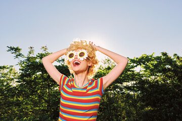 Obraz premium Smiling woman with curly hair wearing flower glasses and a striped shirt in bright sunlight. Outdoor setting conveys playful summer vibes, carefree style, and bold color.