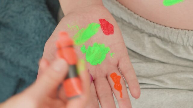 Hand painting on belly, pregnant white lap view of palm resting on lap with neon green and red paint, small hand applying orange and purple dabs, warm domestic setting, relaxed maternity craft