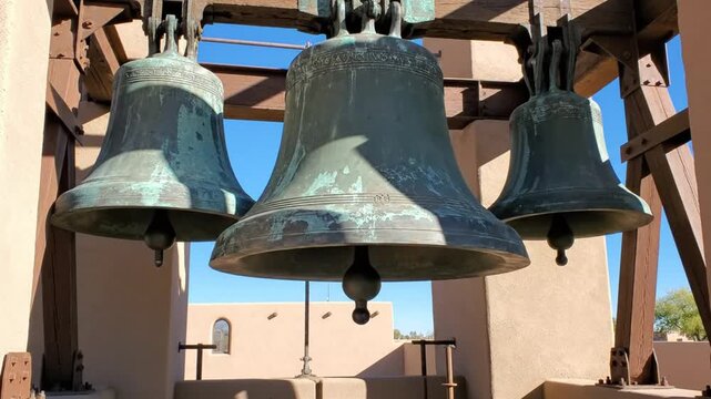 Three large bronze church bells with green patina hanging from wooden supports against a bright blue sky.