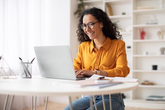 Smiling Latin American Woman Sitting At Table Working On Laptop In Modern Home Office, Enjoying Productive Day, Focusing On Task With Happy Expression. Business Concept.