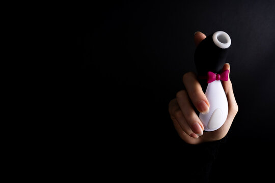 A hand holding a penguin shaped air wave vacuum clitoral stimulator with a pink bow tie against a dark black background, illustrating modern discrete adult pleasure toys.