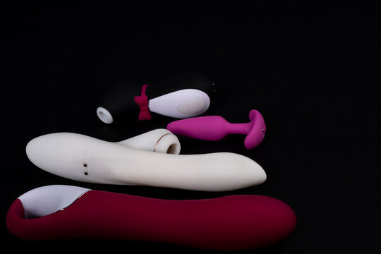 Close up shot of four different adult sex toys including silicone vibrators, clitoral suction stimulators, and a pink butt plug on a dark background.
