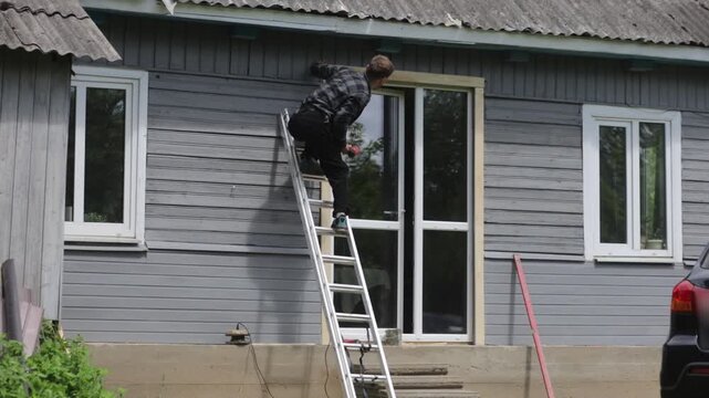 A man is renovating a wooden house, drilling a hole in the frame of a glass door outside the house