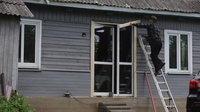 A man is renovating a wooden house, drilling a hole in the frame of a glass door outside the house