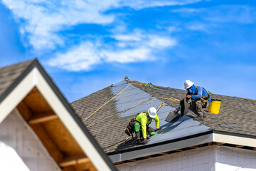 A wide horizontal view of two roofers wearing hard hats and safety harnesses while installing asphalt shingles on the roof of a residential building structure under a bright blue sky with white clouds © Andrei Liashchou