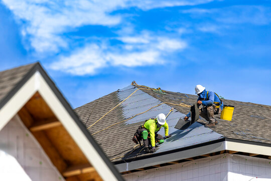 A wide horizontal view of two roofers wearing hard hats and safety harnesses while installing asphalt shingles on the roof of a residential building structure under a bright blue sky with white clouds