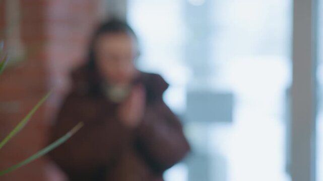 Caucasian woman waits near cafe plant, writer and podcaster pausing with headphones, leaf filled foreground and soft bokeh, glass door blur with snowy street beyond, gentle wave and shy smile, cozy