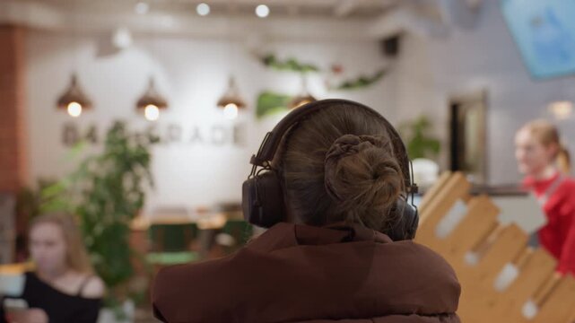 Woman with headphones at cafe counter, back view in brown puffer coat ambient pendant lights and menu screens over a busy counter with barista preparing orders scene shifts as she turns to greet