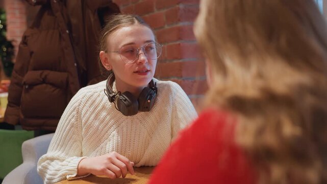 Young white woman listening in cafe wearing chunky sweater and headphones, seated across friend against brick wall, earnest hand gestures and thoughtful expression, warm ambient lighting conveys