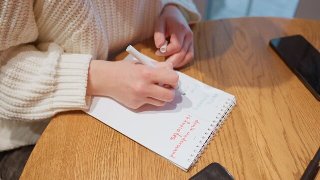 White hands writing in cafe notebook cozy sweater sleeves wooden table smartphone nearby sequence shows drafting notes erasing tapping page putting pen down warm morning mood focused planning