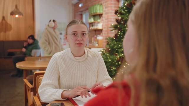 White woman talking in cafe, cream sweater facing friend in red, notebook open on wooden table, decorated tree with lights, warm brick interior, casual holiday conversation, ambient laughter, study