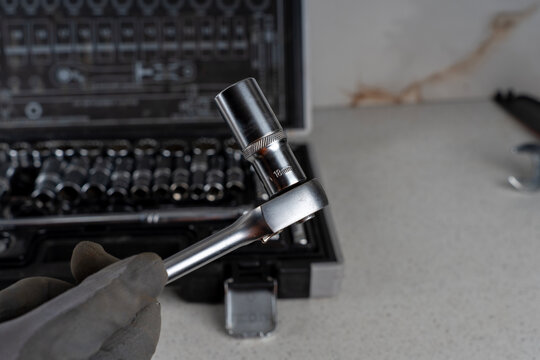 Hand in a protective glove holding a steel ratchet wrench with an attached 18mm socket head, with an open tool case in the blurred background.
