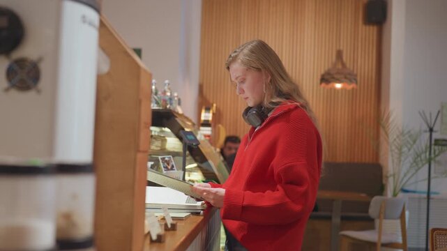 Young woman reading menu at cafe studying pastry display, wearing red sweater with headphones, glass case of baked goods, warm lighting and wood paneling, focused selection and attentive ordering