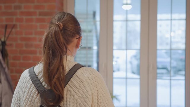 Young white woman entering cafe with backpack and knit sweater, pauses by glass door, scans interior, greets friend, exchanges glance with seated patron, winter light through windows, brick wall