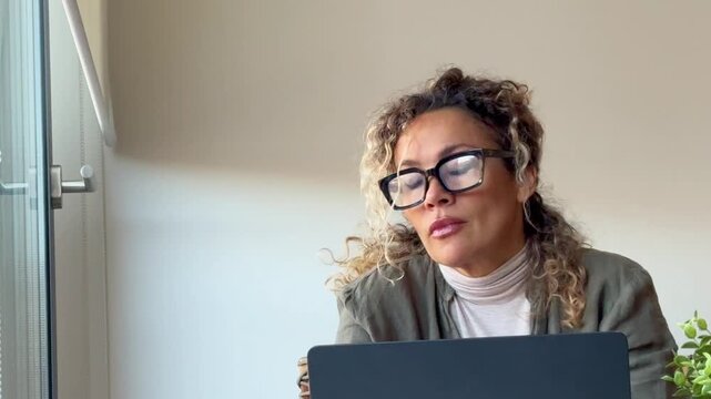 A tired woman with curly hair and glasses yawns and stretches her neck and shoulders while working on her laptop, taking a necessary break from her remote work or studies.