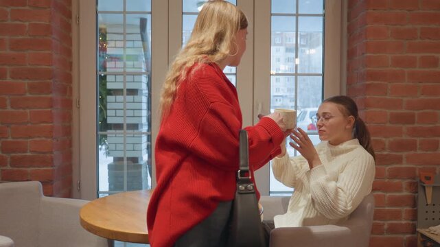Red sweater woman offers coffee in cafe, crossing threshold to table, seated friend in white sweater accepts cup, brief exchange and smiles, window view of snowy street, warm steam, subtle tension