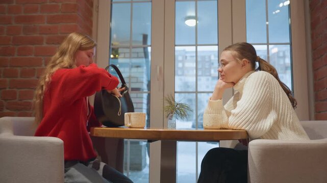 Two white women in cafe conversation and support, red sweater rummages handbag while friend in cream sweater sips coffee and listens thoughtfully, brick wall bistro setting with city window backdrop,