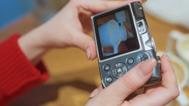 Hands reviewing camera photos of cafe, closeup of white woman hands in red sweater checking digital compact camera screen showing latte mug and foam, intimate cozy tabletop scene, warm ambient light