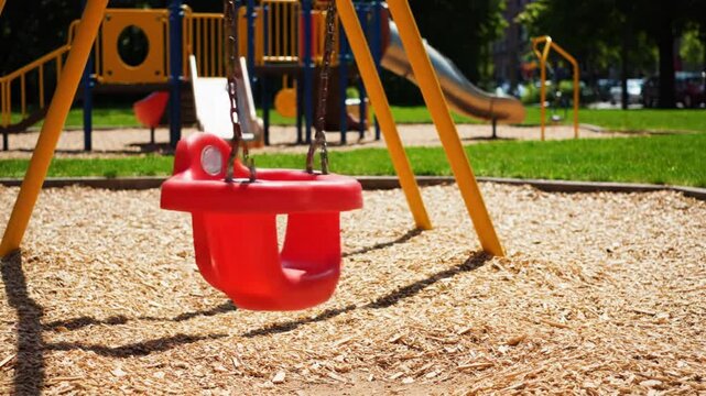 Empty red baby swing gently swinging on a sunny day at a colorful playground