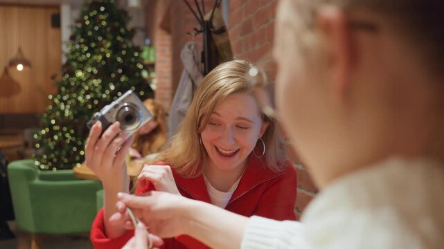 Cafe table woman with camera capturing memory in festive setting, friend reaches to take vintage camera across table, warm brick wall, Christmas tree and green chairs, red sweater, candid laughter