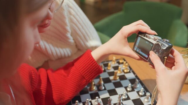 Young white woman in cafe photographing chessboard closeup, intimate hands hold mirrorless camera over chessboard on wooden table, red sweater, green chair, vintage strap, blurred background, warm
