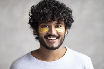 Obraz premium Smiling Young Man With Curly Hair Applying Eye Patches At Home In Relaxing Setting. Enjoying Self-care While Taking Time For Skincare Routine. Focus On Wellness And Personal Care Concept.
