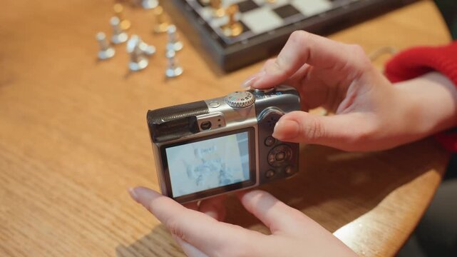 Closeup hands adjusting camera on cafe tabletop, redsleeved person tweaks lcd settings, compact digital camera screen shows framing, scattered chess pieces on wooden surface create cozy atmosphere,