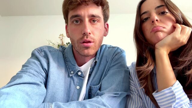 A couple on a video call appears tired and bored, displaying a sense of waiting. Their focused gaze suggests an online interaction or virtual meeting.