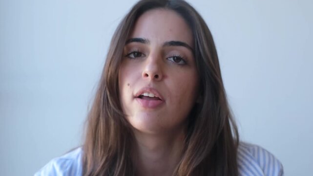 A close-up shot of a young brunette woman as she speaks, closes her eyes in a moment of peace, then smiles gently and opens her eyes again, conveying thoughtful emotions.