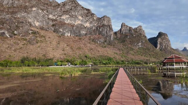 A Pathway to Serenity: A long red wooden walkway stretches across a tranquil lake towards a rustic pavilion nestled at the foot of dramatic, tree-covered mountains under a cloudy sky.