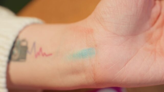 Turquoise glitter swatch on wrist showing finger pressing and blending pigment, camera tattoo with heartbeat line visible near palm, cozy sweater cuff, warm indoor lighting, beauty product tester