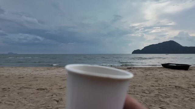 Morning Coffee by the Sea: A close-up of a white coffee cup on a sandy beach with the ocean and cloudy sky in the background, evoking a sense of peaceful morning relaxation.