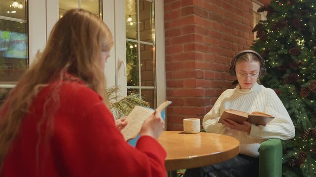 Two women at cafe table with phone and open book, exposed brick and festive lights behind, one scrolling on smartphone while other follows text in book, mugs and plant on table, relaxed urban vibe