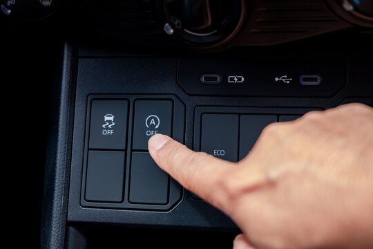 Close up of a driver finger pressing the automatic engine start stop system off button in a modern car interior dashboard panel to disable the fuel saving feature during heavy traffic jam