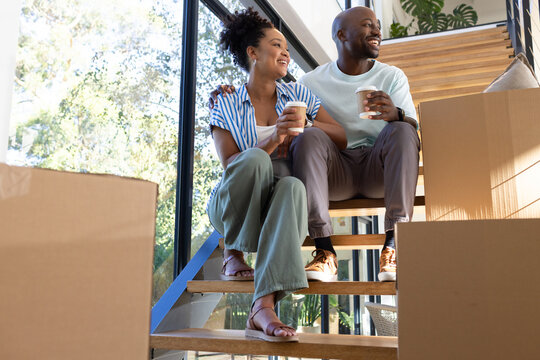 African American couple sitting on wooden stairs at home, holding coffee cups near boxes