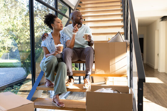 African American couple sitting on wooden stairs in home, holding cups, pointing at moving boxes
