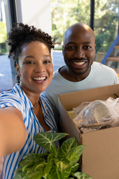 African American couple smiling, taking selfie at foyer holding plant, box, smartphone