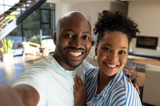 African American couple smiling, posing for selfie in modern living room with phone, striped shirt