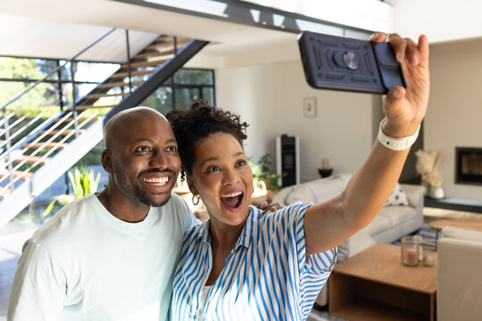 African American couple posing and smiling, holding smartphone in modern living room near staircase