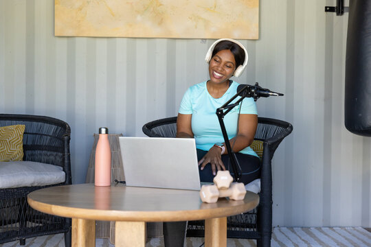 African woman sitting in nook wearing teal tee white headset recording podcast with laptop mic