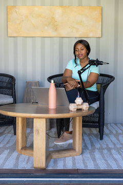Adult African woman sitting in wicker chair recording with mic and laptop in studio, copy space